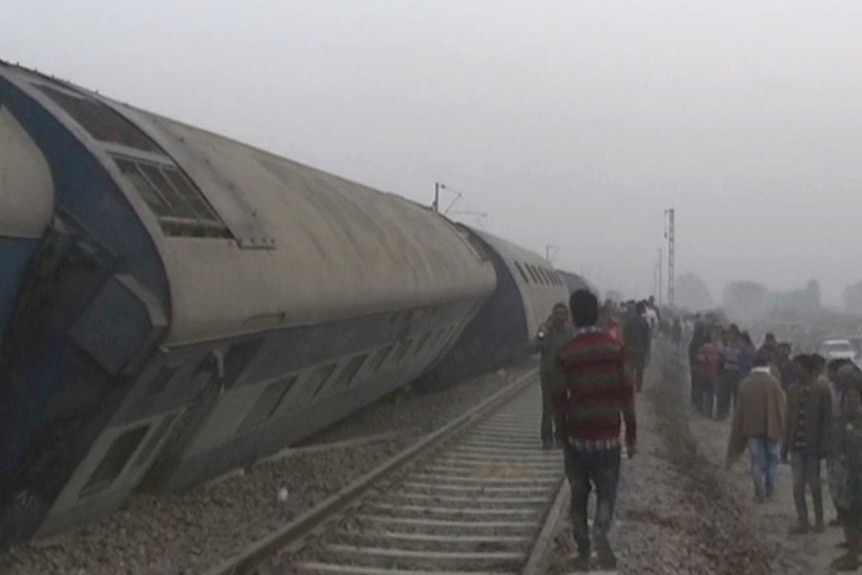 People gather at the site of a train accident near Pukhrayan, a train is shown leaning on its side along the track