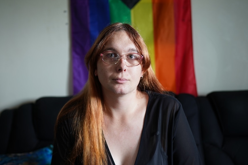 A woman sits in front of a rainbow flag which is hanging on a wall.
