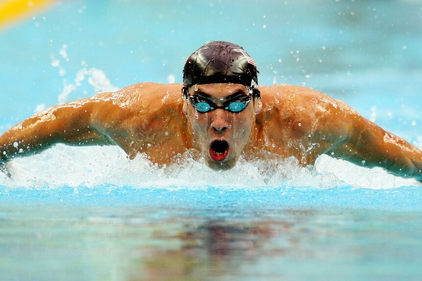 Michael Phelps in a pool swimming wearing goggles and a swimming cap, arms outstretched while doing Butterfly