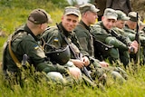 A row of young men in military uniforms holding rifles sit together in tall grass, smiling and talking 
