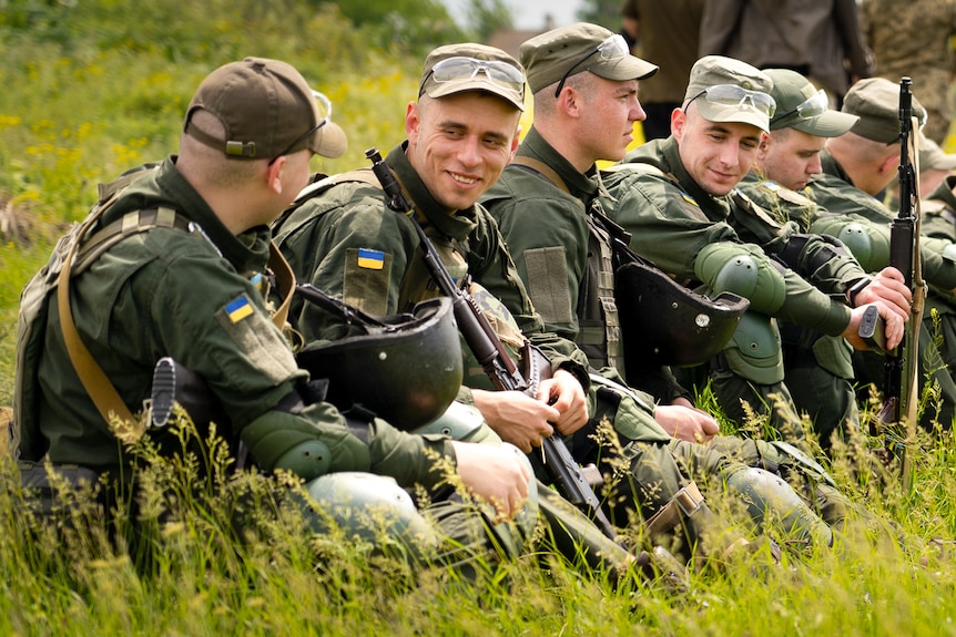 A row of young men in military uniforms holding rifles sit together in tall grass, smiling and talking 