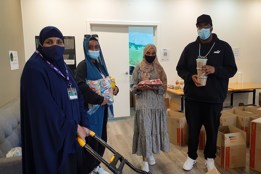 Three women in hijab and face masks and a man stand with cartons. 