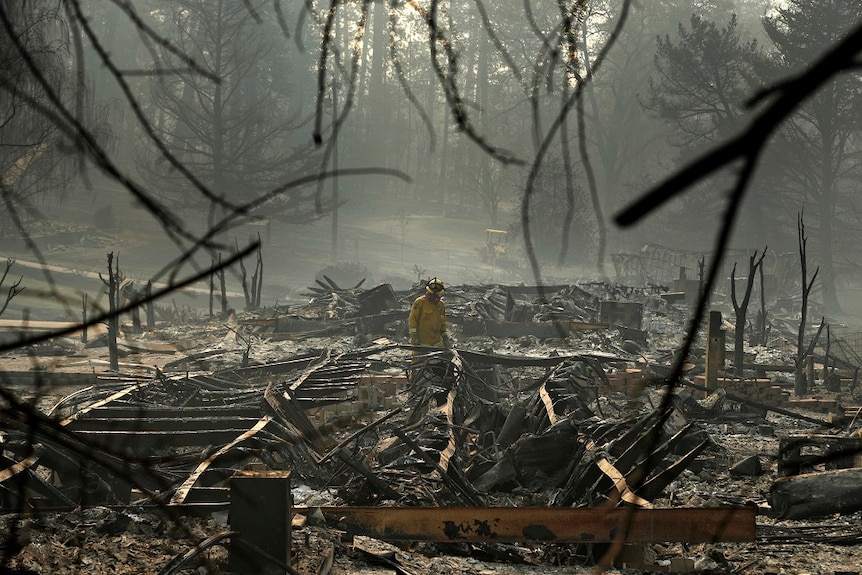 A firefighter works through the collapsed frames and charred wreckage of a trailer home destroyed by fire.