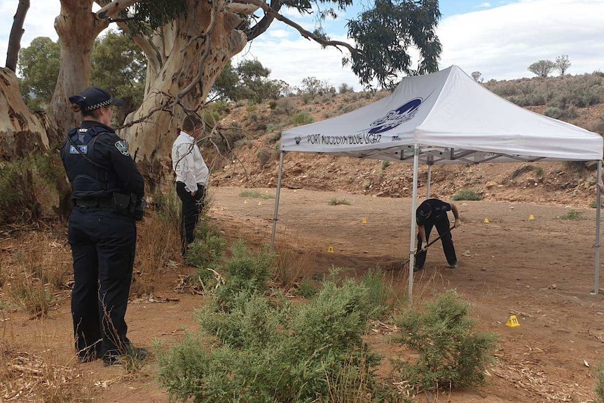 Several police officers stand and watch another police officer digging in dirt under a marquee.