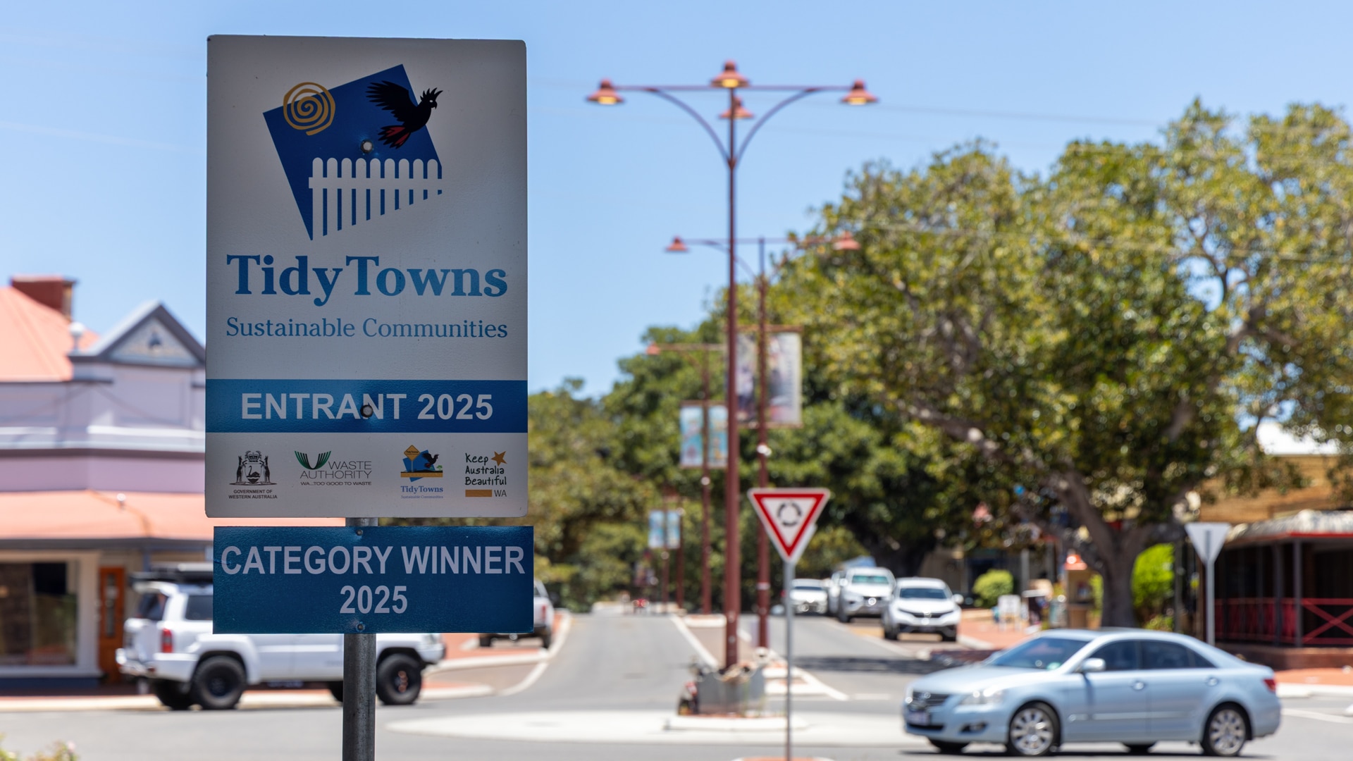 Um sinal que diz "Vencedor da categoria Tidy Towns 2025" ao lado de uma estrada enquanto os carros passam ao fundo.