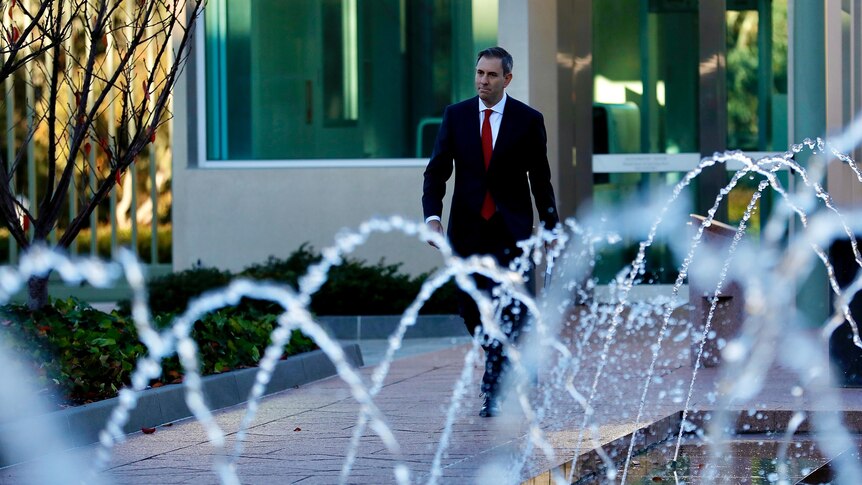 A middle-aged man in a suit walks down a path in a nice garden with sprinklers on in the foreground.