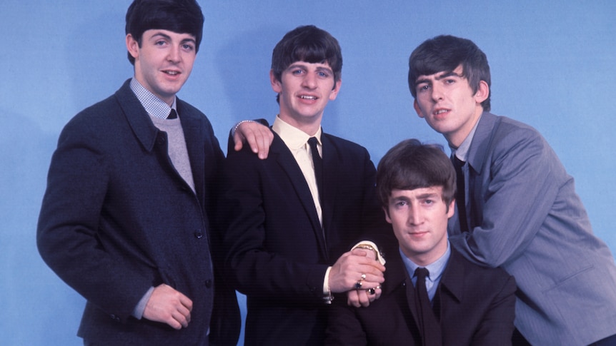 The Beatles at a photoshoot in 1963. They wear suits and stand before a blue background.