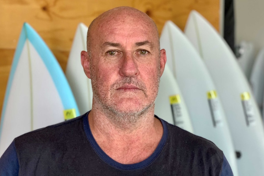 A bald, middle-aged man in a black T-shirt looks at the camera solemnly with five surfboards behind.