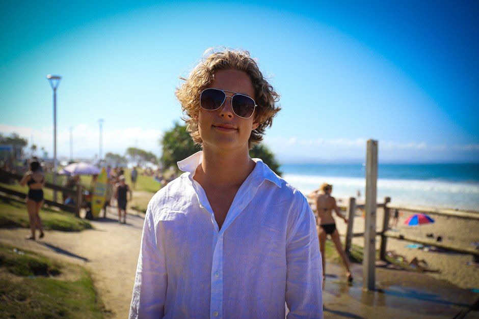 A young man wearing sunglasses and a white cotton shirt standson a path with the beach and ocean in the background.