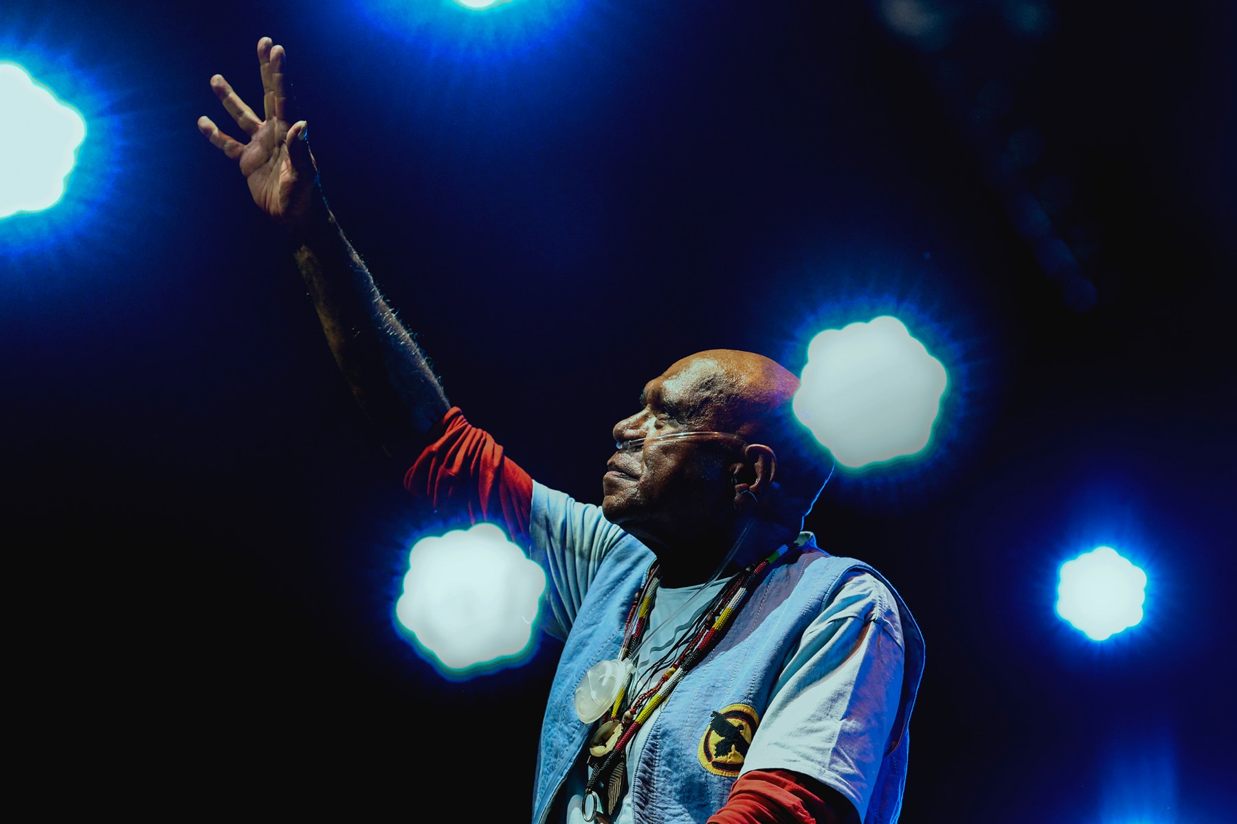 A bald man in a blue jacket over a red shirt sits on a stage with one arm raised in the air.