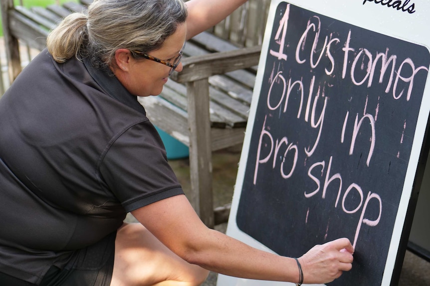 A worker at the Marrickville Golf Club writes a sign preventing more than one person in the shop at a time.