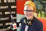 Woman wearing glasses and with headphones around neck in front of microphone in radio studio.