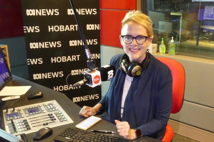 Woman wearing glasses and with headphones around neck in front of microphone in radio studio.