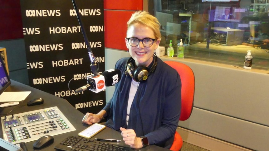 Woman wearing glasses and with headphones around neck in front of microphone in radio studio.