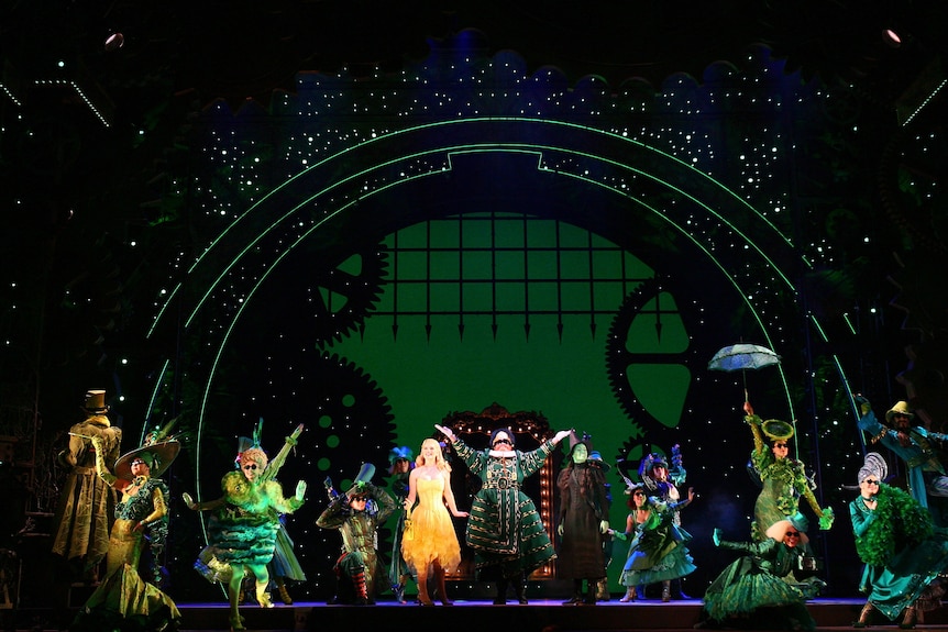 On stage a blonde white woman in champagne dress sings with green-skinned woman in black witch's cloak on elaborate green set.