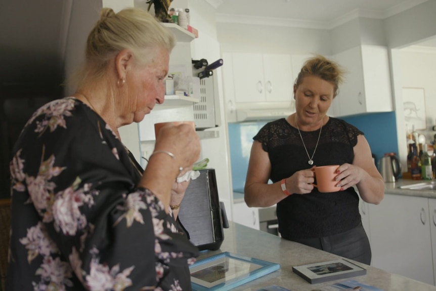 Women stand in a kitchen looking at photos.