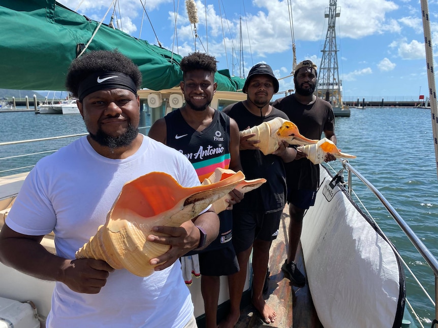 Four Indigenous men stand on a ship holding trumpet shells called Maber Sor.