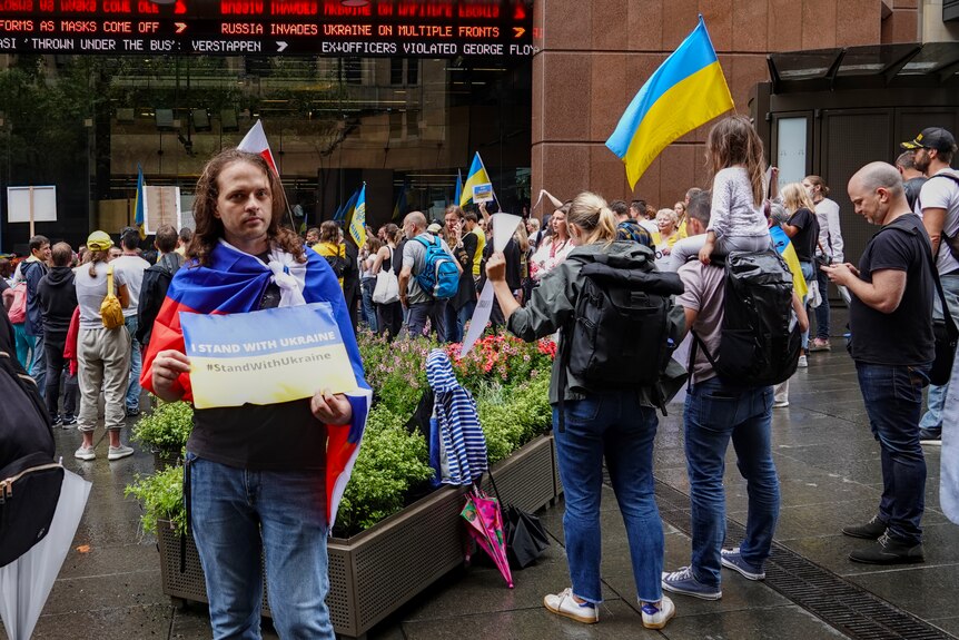 People hold flags at a street protest while one man faces the camera with a sign "I stand with Ukraine".