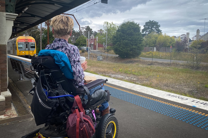 Woman sitting in a wheelchair faces towards an oncoming train on the platform of a station