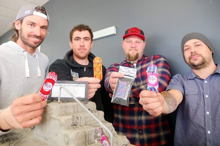 A close-up of four guys holding miniature skateboards out in front with a mini skateboard ramp in the foreground.