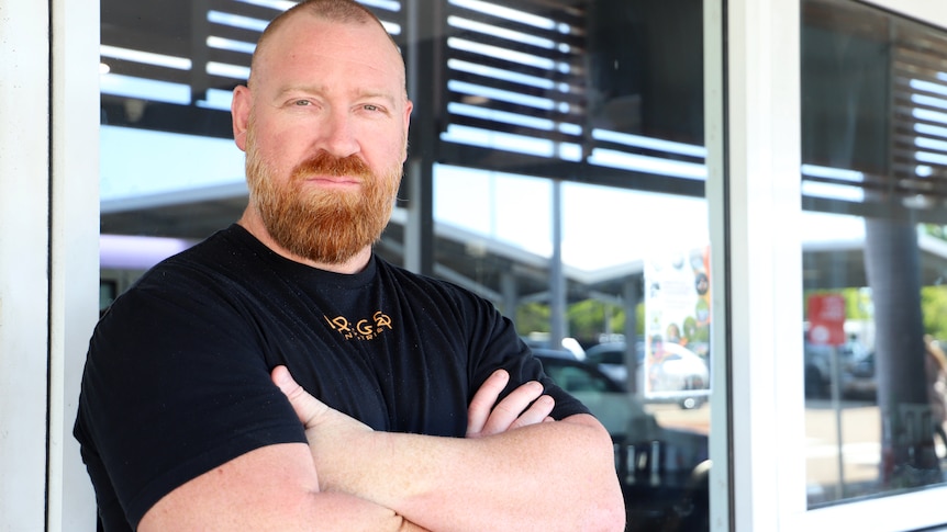 Man in black shirt with red beard stand with arms crossed in front of shop front