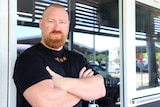 Man in black shirt with red beard stand with arms crossed in front of shop front