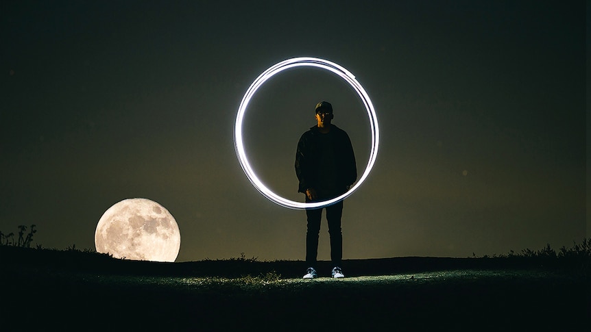 A man stands in front of the full moon making a circle by spinning a light through the air.