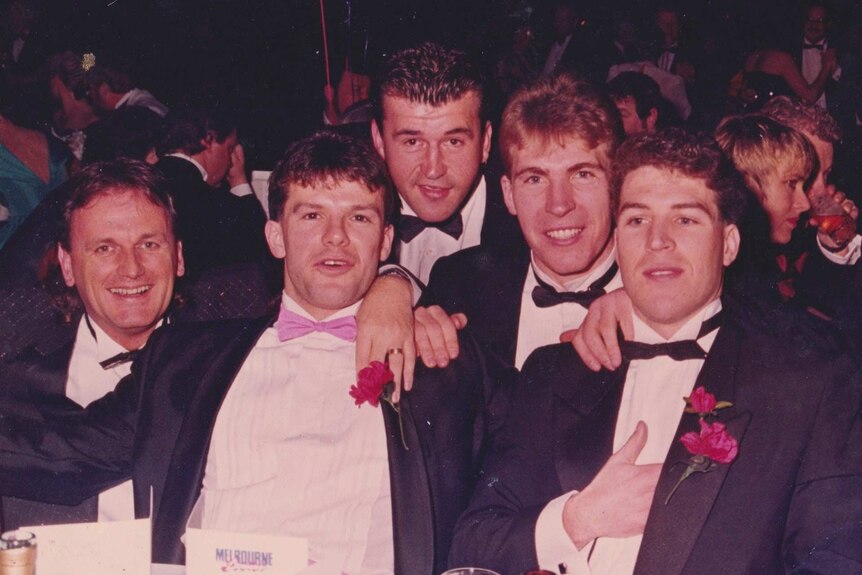 Five men in suits and bow ties sit around a table and look at the camera.