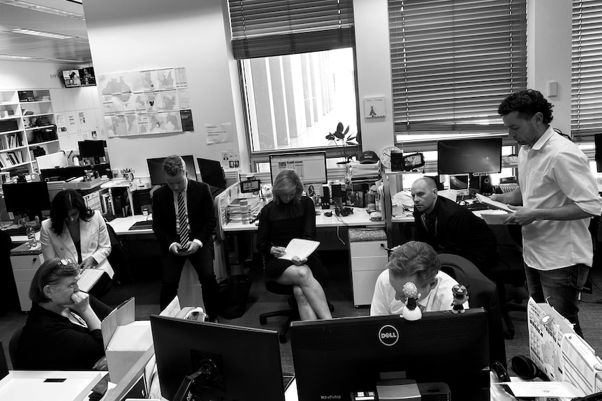 Black and white photo of people in an office listening to a speaker phone and looking concerned.