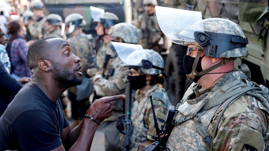 An African American man talks to a member of the National Guard wearing a helmet and face mask.