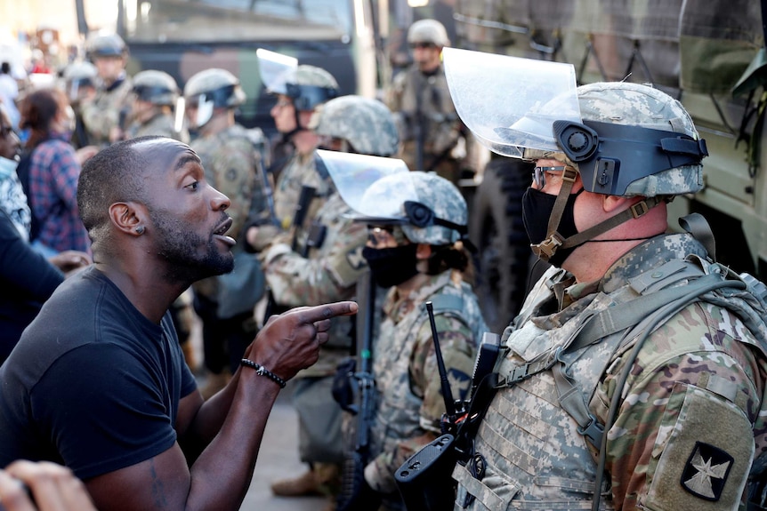 An African American man talks to a member of the National Guard wearing a helmet and face mask.