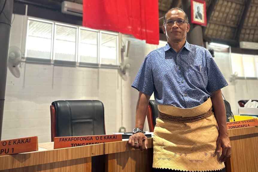 A man in a shirt and traditional woven Tongan ta'ovala leans on his desk in front of the Tongan flag.