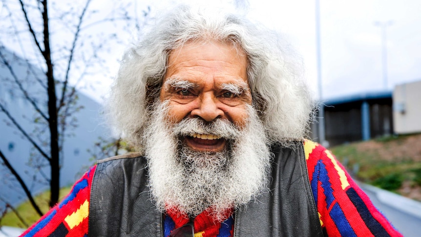 Uncle Jack Charles smiles, dressed in a colourful jumper as he stands outside.