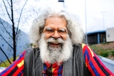 Uncle Jack Charles smiles, dressed in a colourful jumper as he stands outside.
