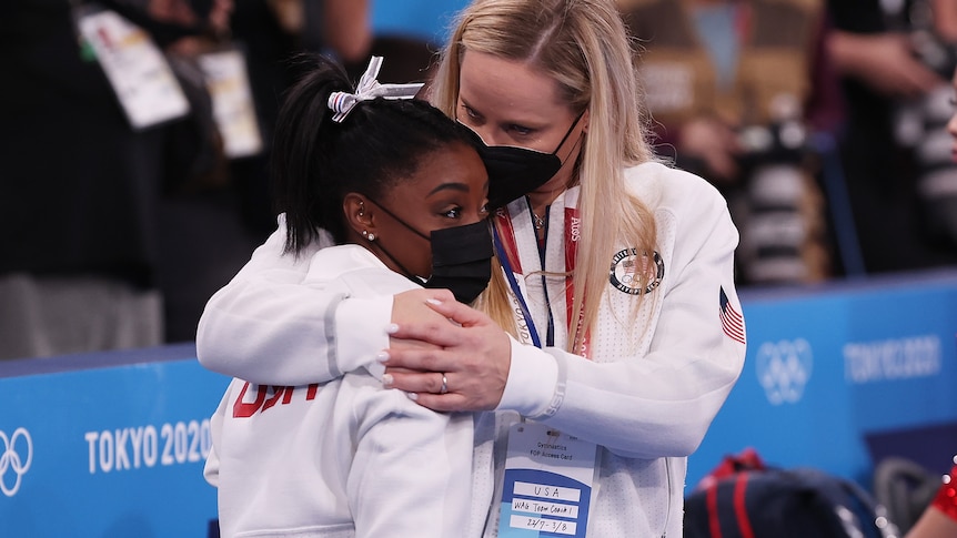 A young woman is hugged by another. They wear matching tracksuits.