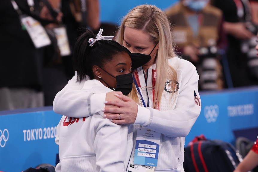 A young woman is hugged by another. They wear matching tracksuits.