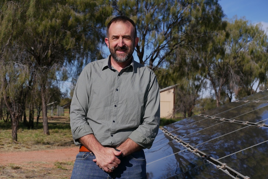 A man half sits on a solar panel, holding his hands in front of him. He smiles into the sun.