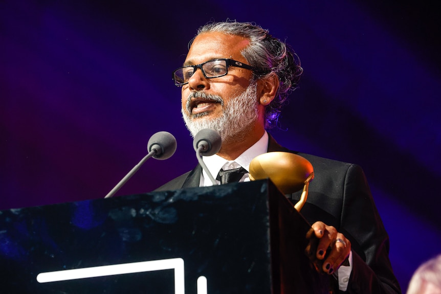 47-year-old Sri Lankan man with grey beard and long hair pulled back, wearing glasses, speaking into lectern-mounted mics.