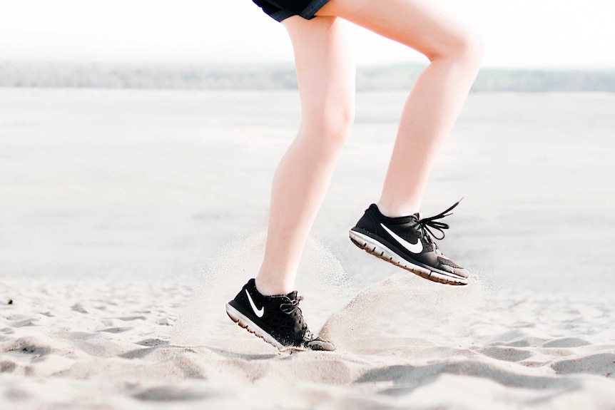 A person wearing sneakers runs on the spot on soft beach sand.