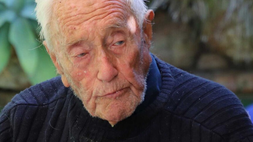 A very old man in a navy jumper sits at an outside table with glasses of wine in front of him.
