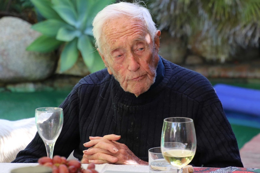 A very old man in a navy jumper sits at an outside table with glasses of wine in front of him.