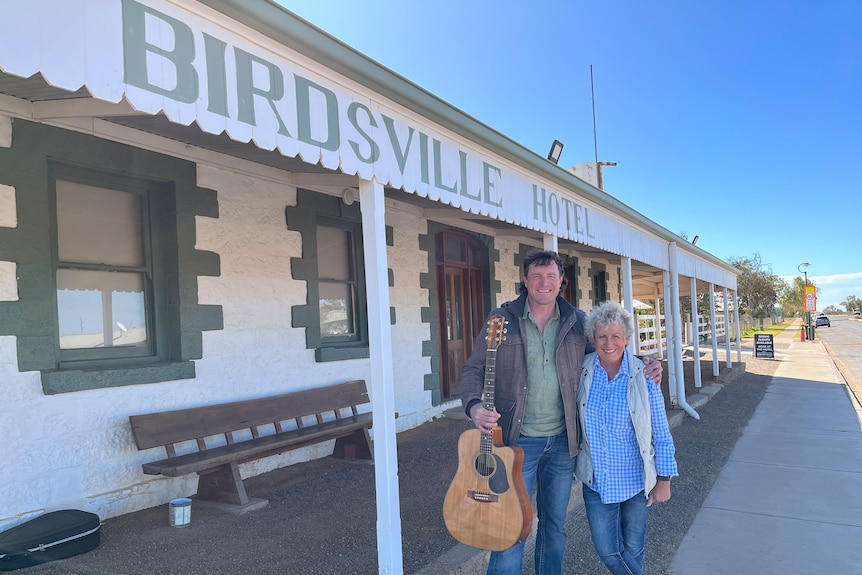 Man holding guitar with arm around woman standing outside old pub called Birdsville Hotel. 