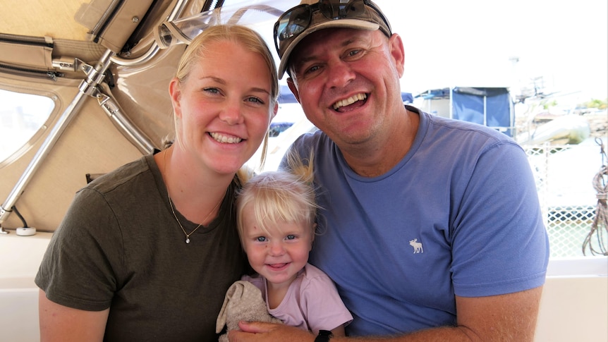 A blonde blue-eyed mother, her one-year-old daughter with pig tails, and her husband who is wearing a cap.   