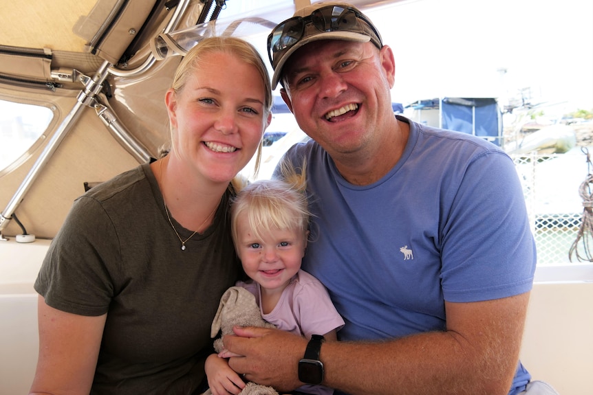 A blonde blue-eyed mother, her one-year-old daughter with pig tails, and her husband who is wearing a cap.   