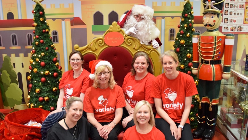 A group of women wearing red Variety shirts smile, crouching around an ornate gold chair. Santa is standing behind the chair.