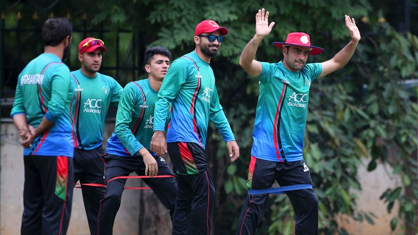 Afghan cricket team members in green team uniforms standing on a training pitch with one to the fore with arms raised.