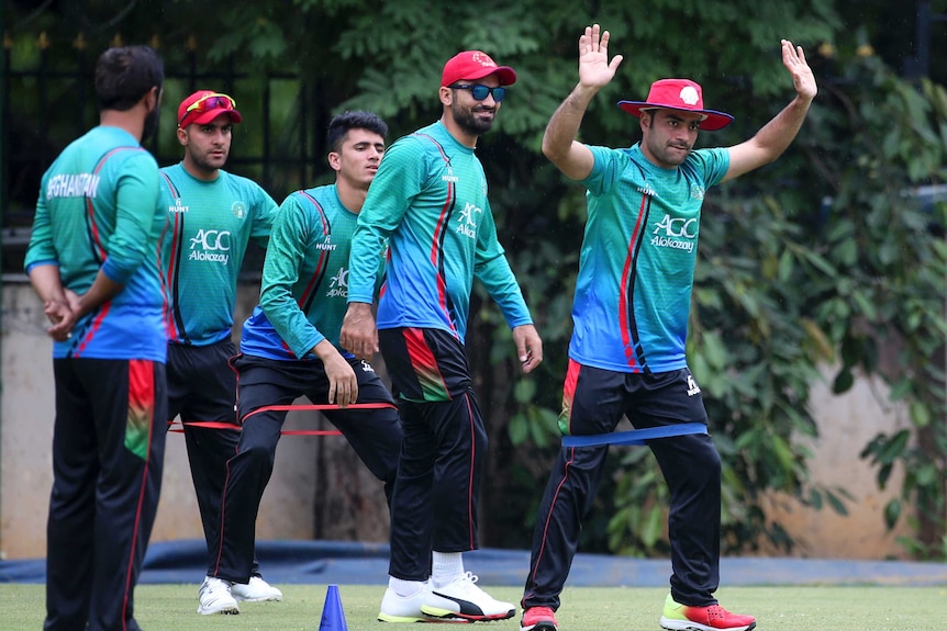 Afghan cricket team members in green team uniforms standing on a training pitch with one to the fore with arms raised.