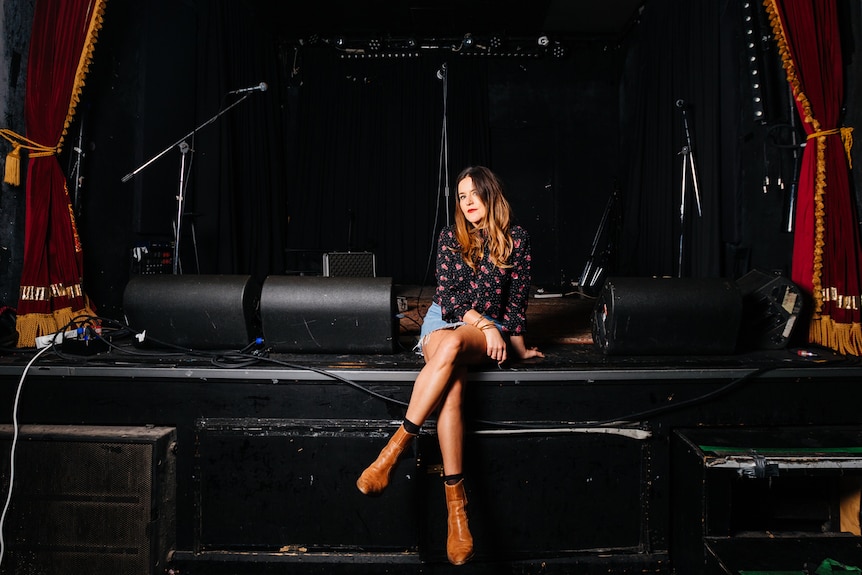 Jess Beston sits on the edge of an empty stage, legs crossed, with microphone stands and speakers behind her