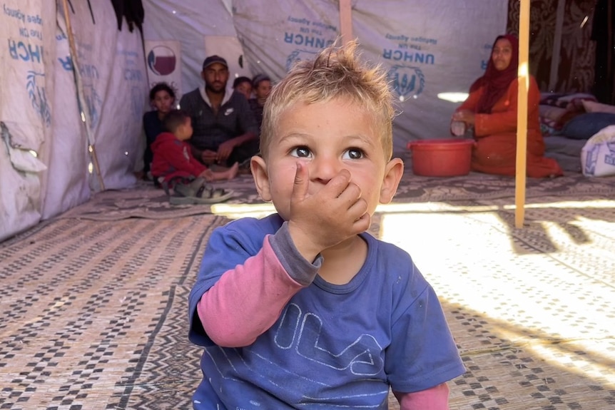 A toddler sits on the ground inside a tent, looking on, with his hand over his mouth, while several relatives sit behind