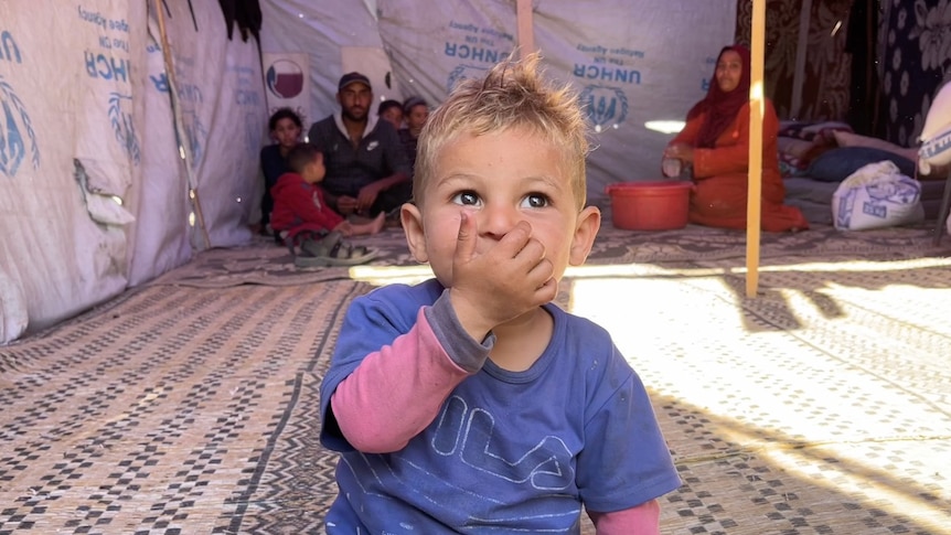 A toddler sits on the ground inside a tent, looking on, with his hand over his mouth, while several relatives sit behind